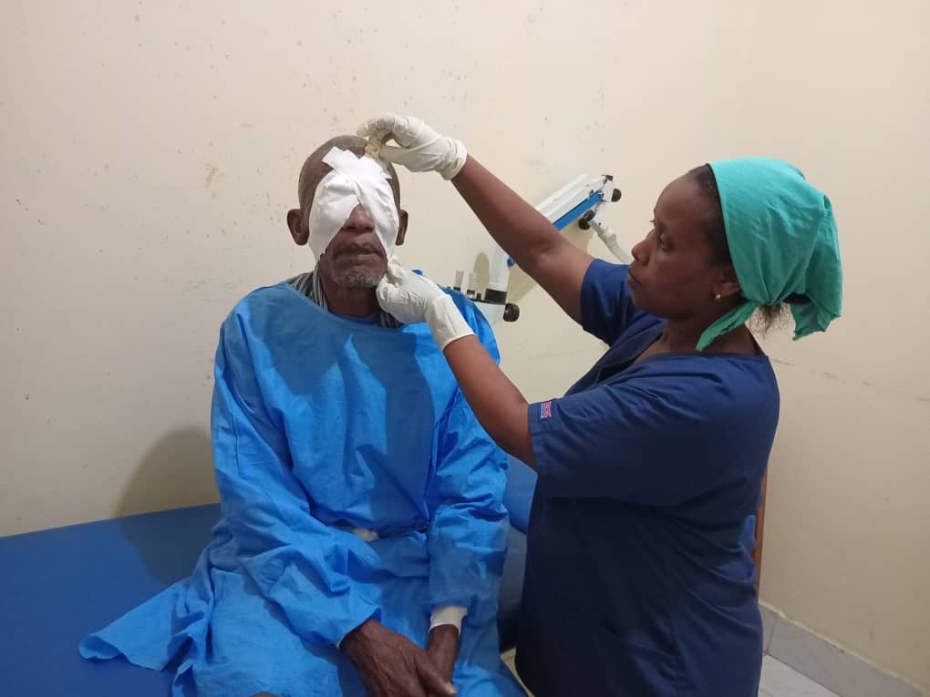 A picture of a nurse helping a man after an eye surgery operation. WOman is putting what looks like a plaster on his two eyes