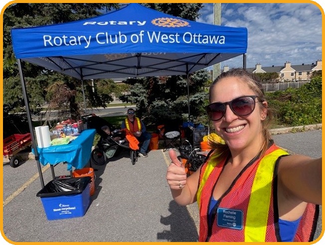 A volunteer gives a thumbs up in front of the blue Rotary Club of West Ottawa tent, with supply tables, orange buckets, recycling bins and a wagon behind.