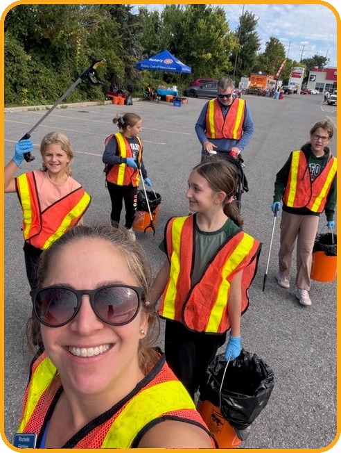 Five young volunteers in high visibility vests hold litter pickers, buckets and garbage bags in a parking lot, with an adult volunteer and the Rotary tent behind them.