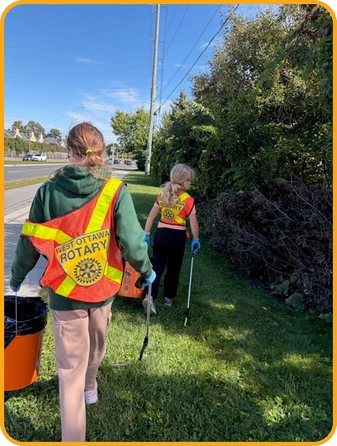 Two young volunteers in West Ottawa Rotary vests walk along a grassy verge beside a road, carrying litter pickers, an orange bucket and a garbage bag, with bushes and trees beside them.