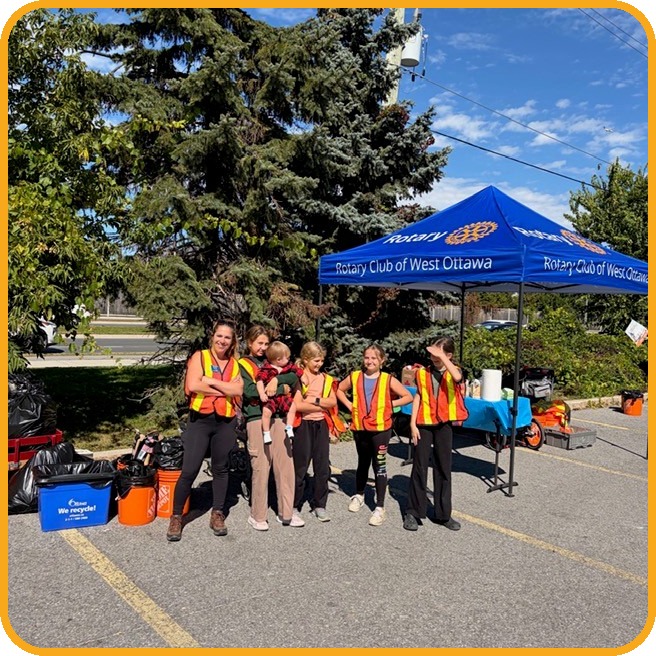 Six Rotary Club of West Ottawa volunteers, most of them children, stand in high visibility vests beside filled garbage bags and orange buckets, in front of the club's blue Rotary Club of West Ottawa tent.