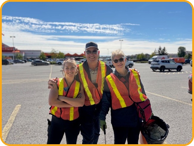 Three adult Rotary Club of West Ottawa members in high visibility vests stand together in a parking lot, one holding a filled garbage bag.