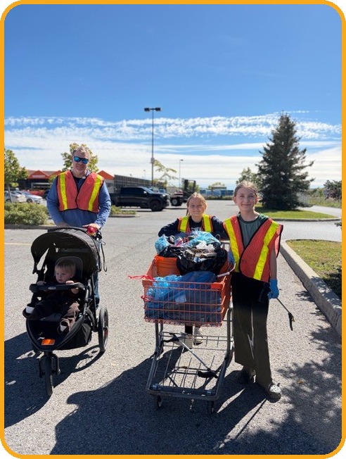 Two young volunteers and an adult with a stroller stand behind a shopping cart heaped with collected litter in a parking lot.
