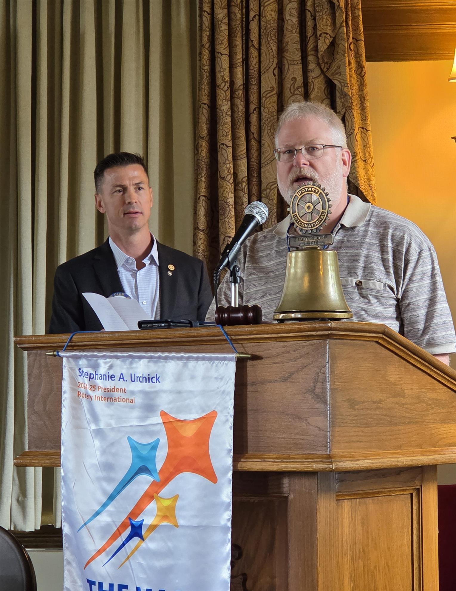 A person standing at a podium with a bell and a person standing behind himAI-generated content may be incorrect.