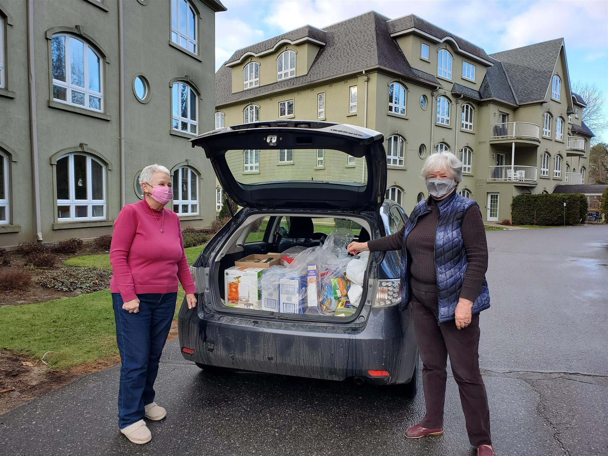 A photo of two women standing beside the open trunk of a car. The trunk is filled with items for the food bank.