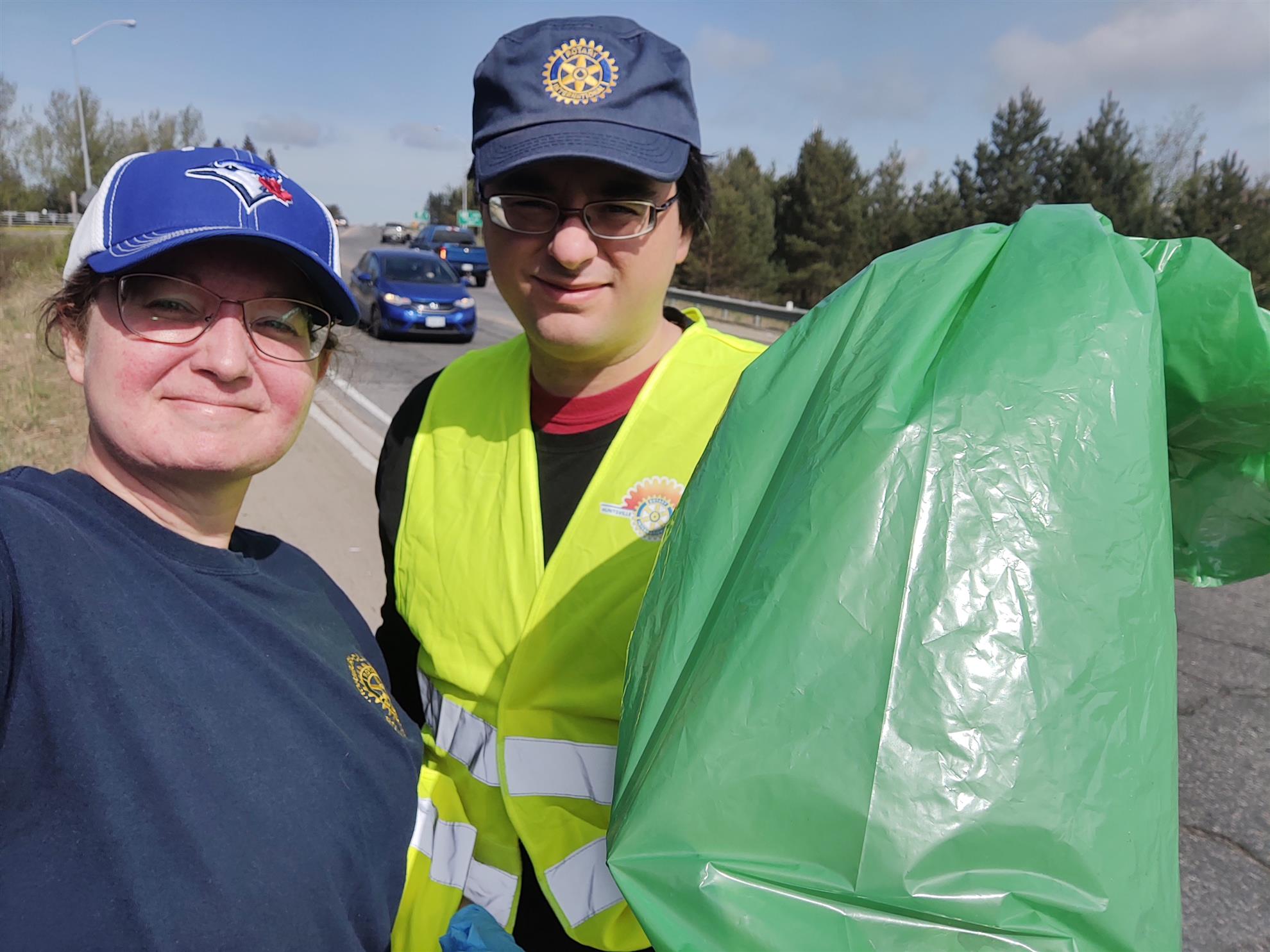 A woman in a dark blue shirt with Rotary logo and man in yellow reflective vest with blue cap with Rotary logo stand by a roadway with a green trash bag.