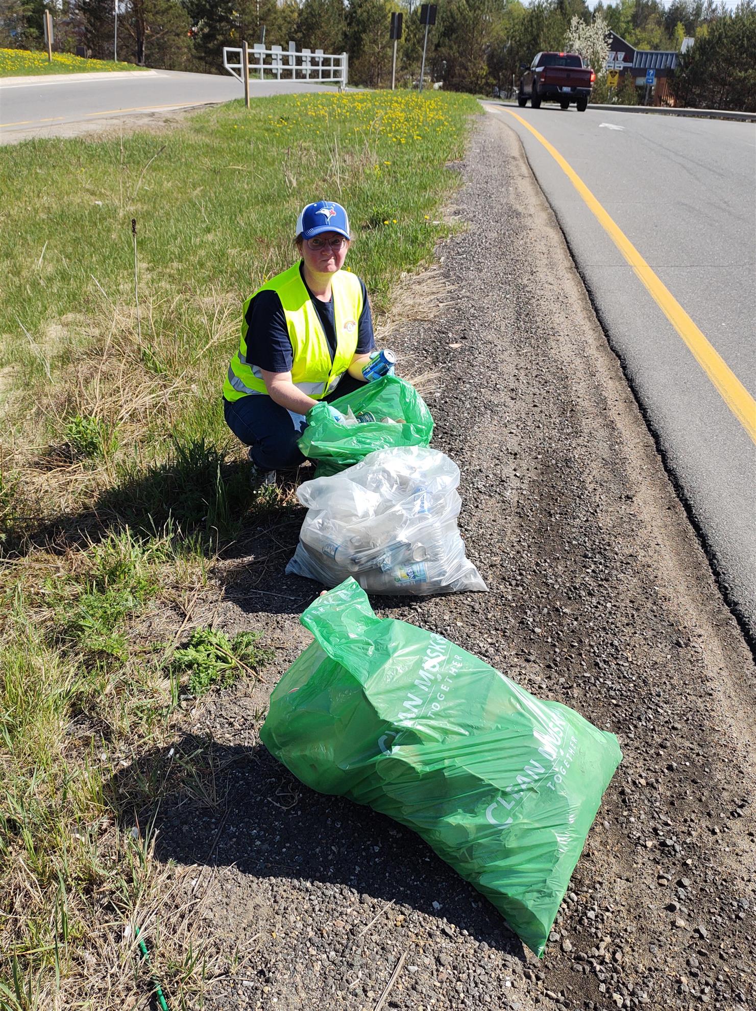 A woman frowns while crouching beside 3 bags of litter at a roadside.