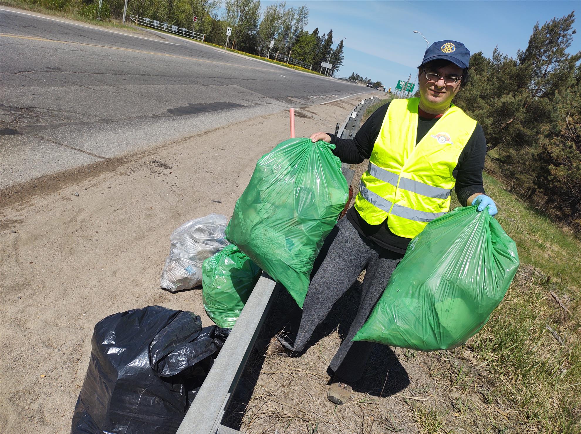 A man in a yellow reflective vest and blue hat with yellow Rotary logo stands near a roadside guardrail holding 2 bags of trash with 3 more beside him.