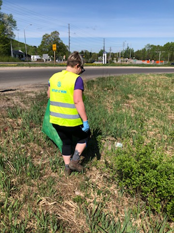 A woman in a yellow reflective vest holds a green trash bag while walking on grass beside a roadway.