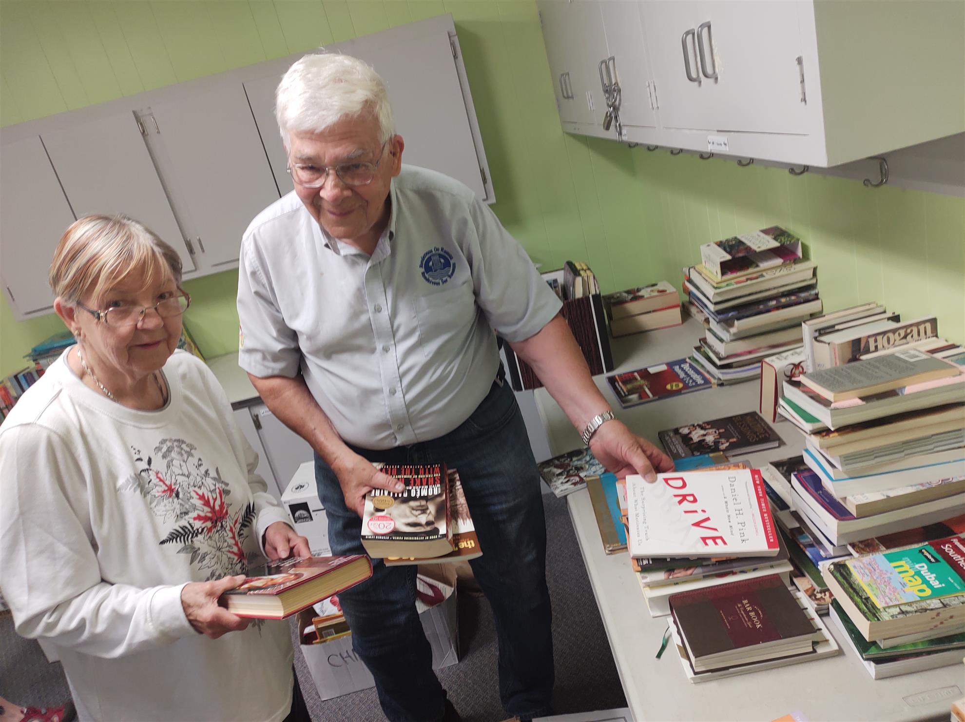 An older couple smiles for the camera while sorting a table filled with books.