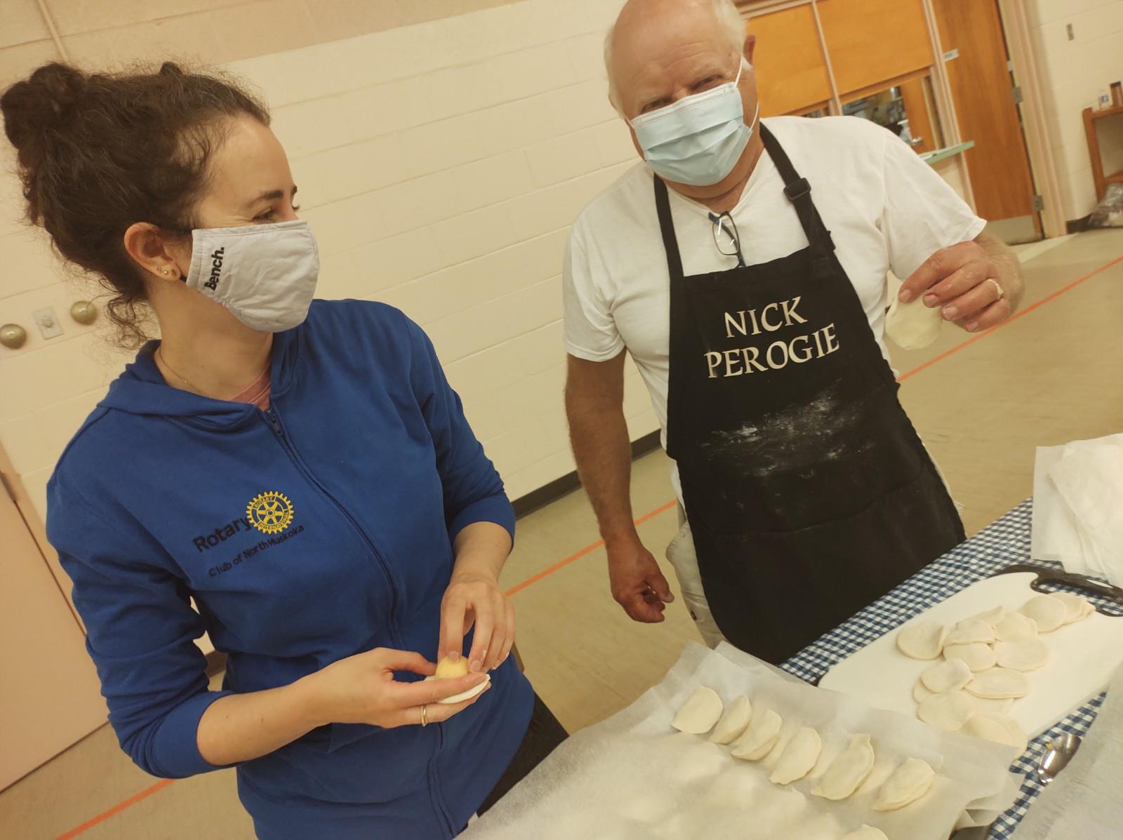 A woman and a man, both wearing face coverings, smile while making perogies.