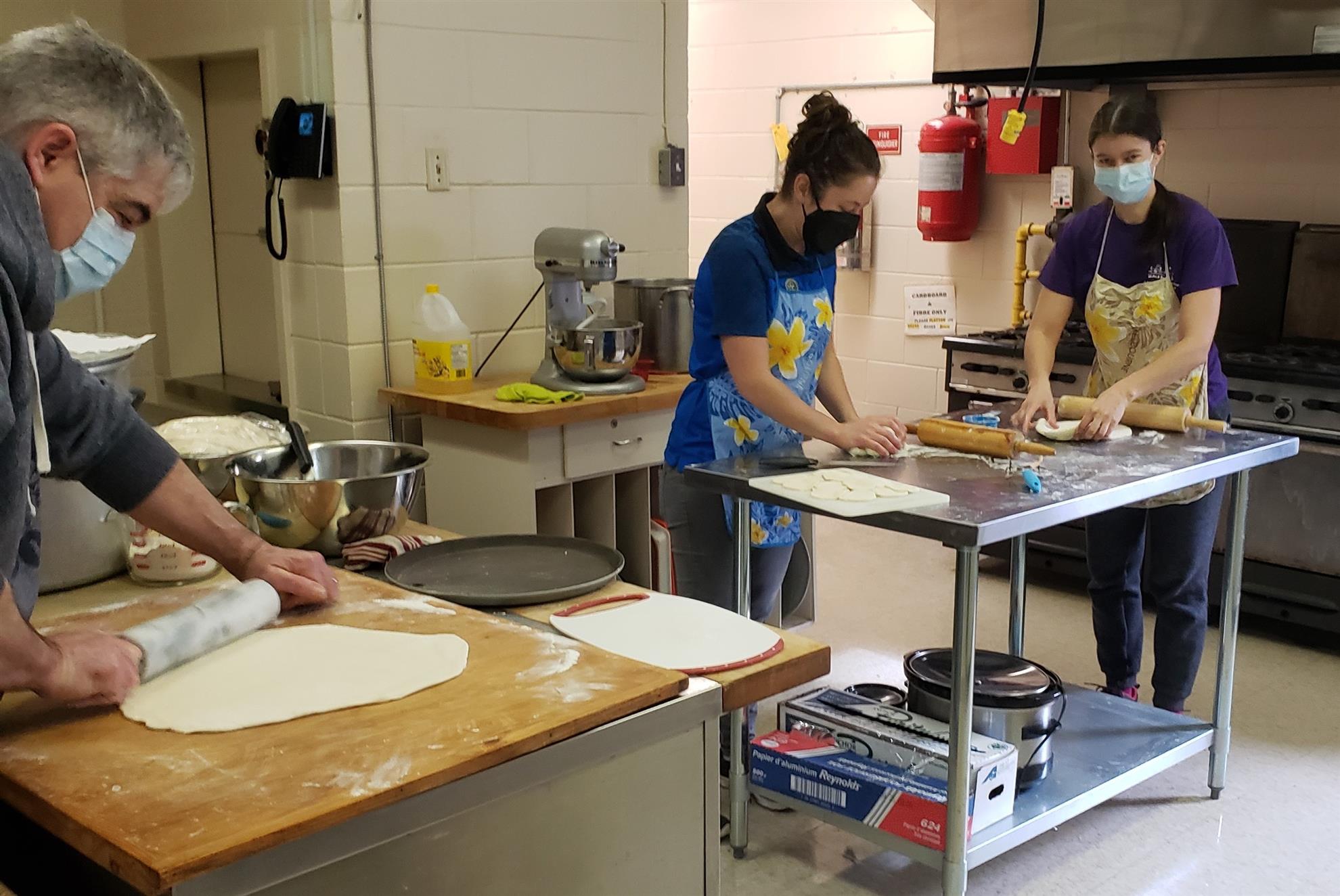 A photo of 3 people, who wear masks and aprons, in a commercial kitchen, rolling and cutting out dough for perogies