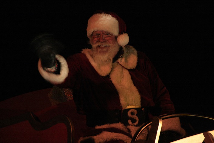 Santa Claus, dressed in a red and white hat and coat, waves to the crowd during a nighttime parade. 