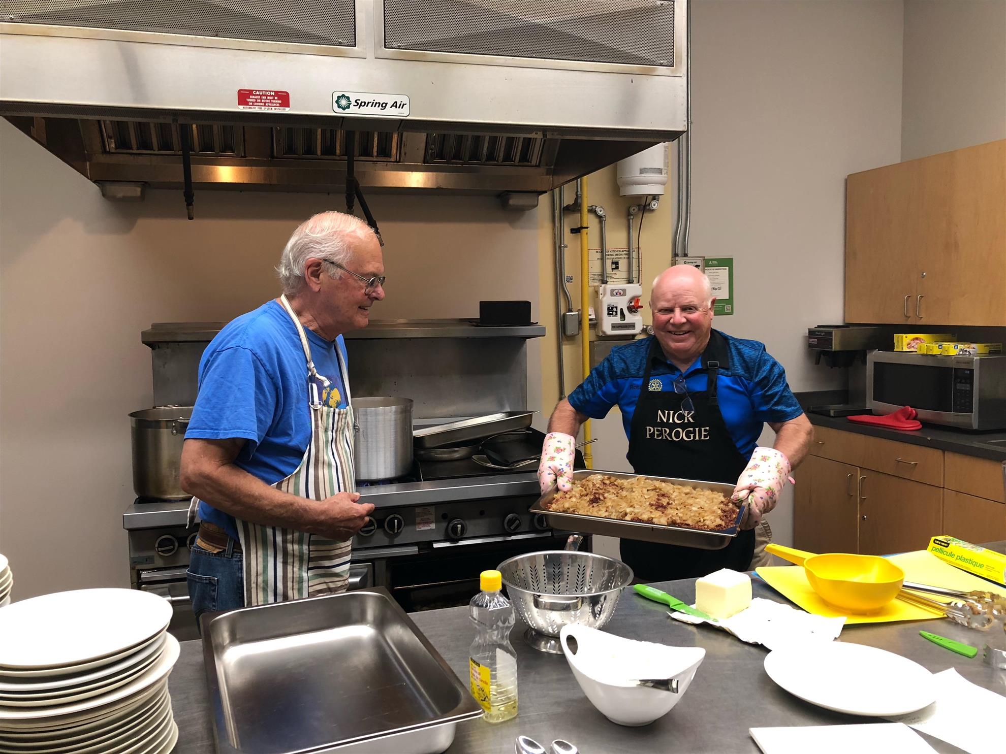 2 smiling men wearing aprons in a commercial kitchen. 1 man is holding a large tray of apple crisp.