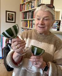 A person holding two glasses of green-colored cocktails, standing in a book-filled room.AI-generated content may be incorrect.