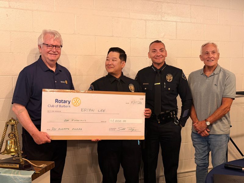 Four individuals stand indoors against a light-colored brick wall during a Rotary Club presentation. Two uniformed police officers stand in the center holding a large ceremonial check from the Rotary Club of Burbank made out to Brian Lee for $1,000 as the Rex Andrews Award. One person in a Rotary polo shirt stands beside the recipients, while another stands at the opposite end of the group. A Rotary bell is visible on a table in the foreground.