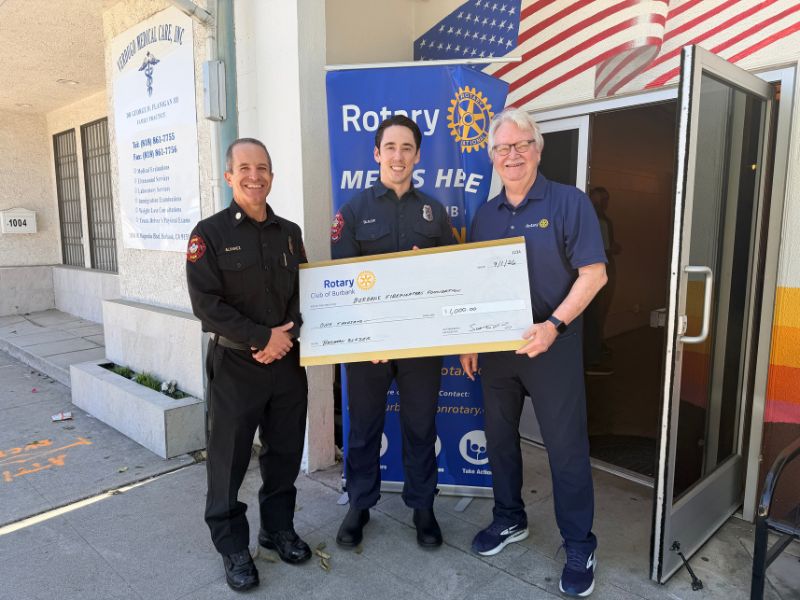 Alt text: Three individuals stand outside a building entrance in front of a Rotary membership banner and an American flag mural. Two members of the Burbank Fire Department and a Rotary Club representative hold a large ceremonial check for $1,000 made out to the Burbank Firefighters Foundation. The group poses on the sidewalk beside an open doorway on a sunny day.