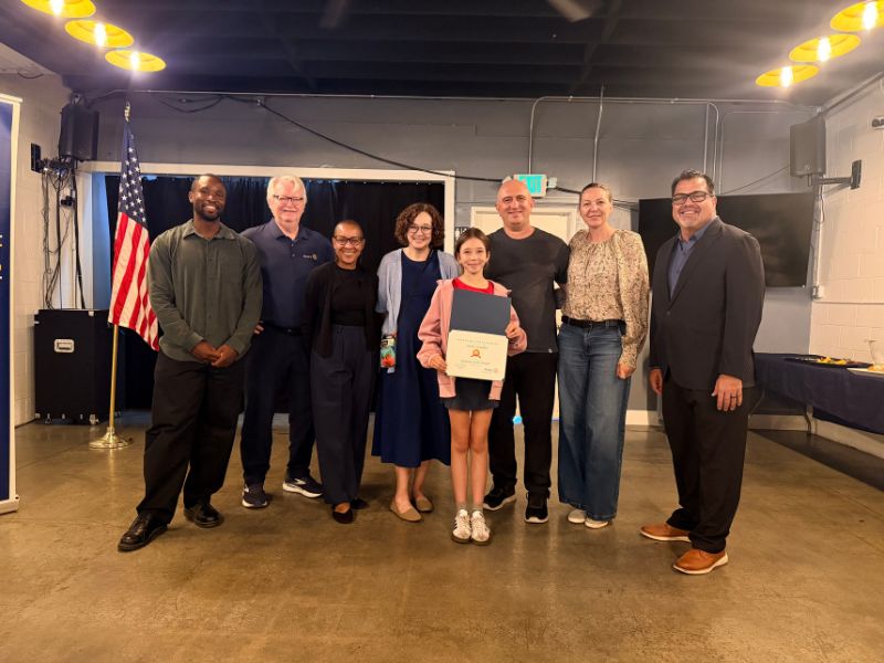 Eight people stand together indoors during a student recognition ceremony. At the center, a student holds a framed certificate while surrounded by family members, school representatives, and Rotary Club members. The group poses in a meeting room with polished concrete floors, an American flag in the background, and overhead pendant lights. Tables line the sides of the room as the student is recognized for an award.