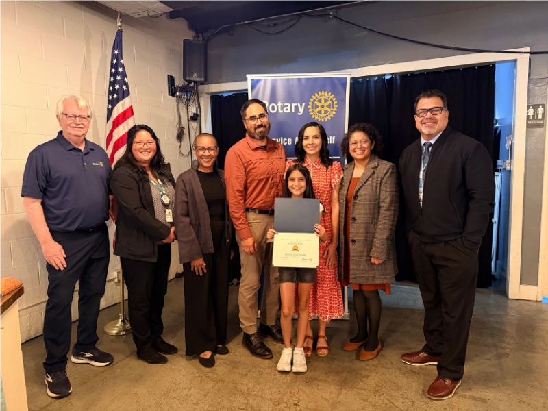 Student of the Month award recipient stands at the center of a group photo holding a certificate, surrounded by family members, school representatives, and Burbank Noon Rotary Club members. The group poses in front of a Rotary banner and an American flag during an award presentation ceremony recognizing the student's achievements.