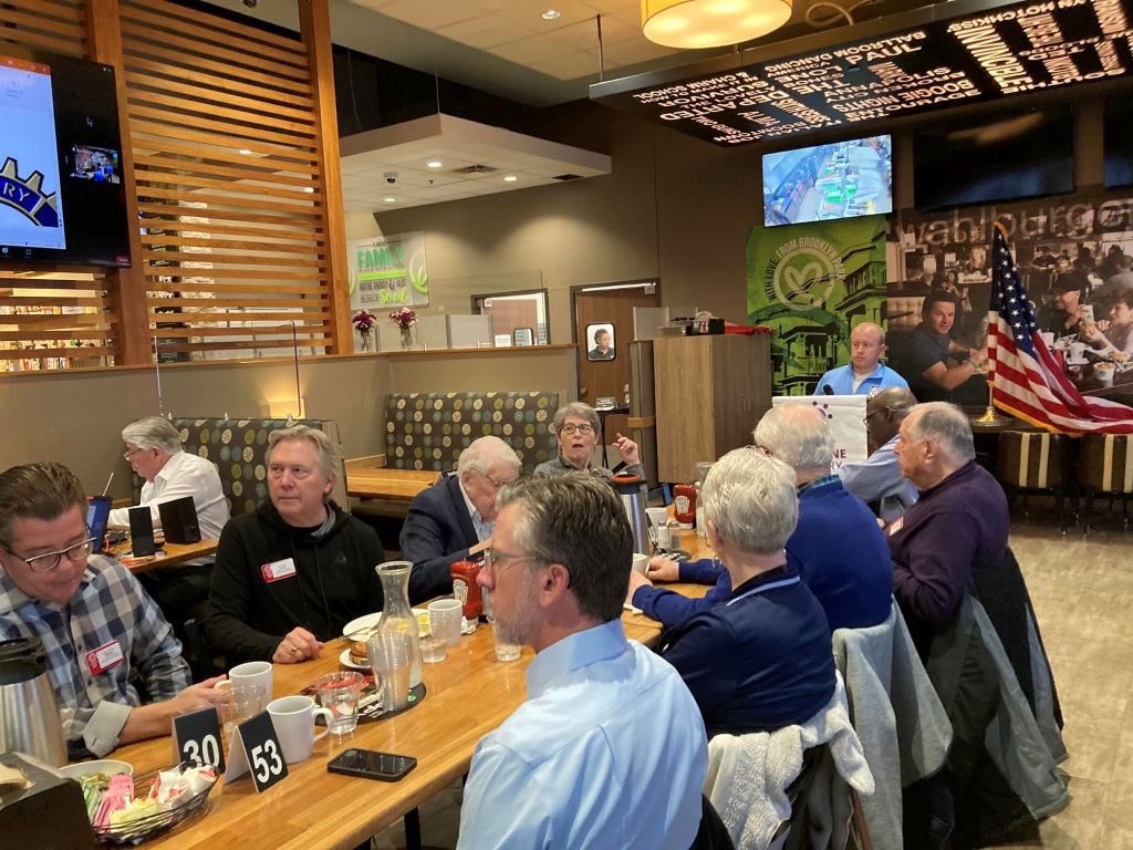 A three-quarters view of people sitting at a long table in the HyVee Wahlberger restaurant. Carl Lottman, the Vocational Services manager of the rotary club, stands at the podium.