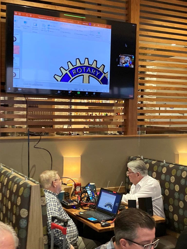 Two rotarians converse at a booth in the HyVee Wahlberger restaurant. They have laptops, tablets, and speakers connected to flat-screen monitor hung on the wall curtains.