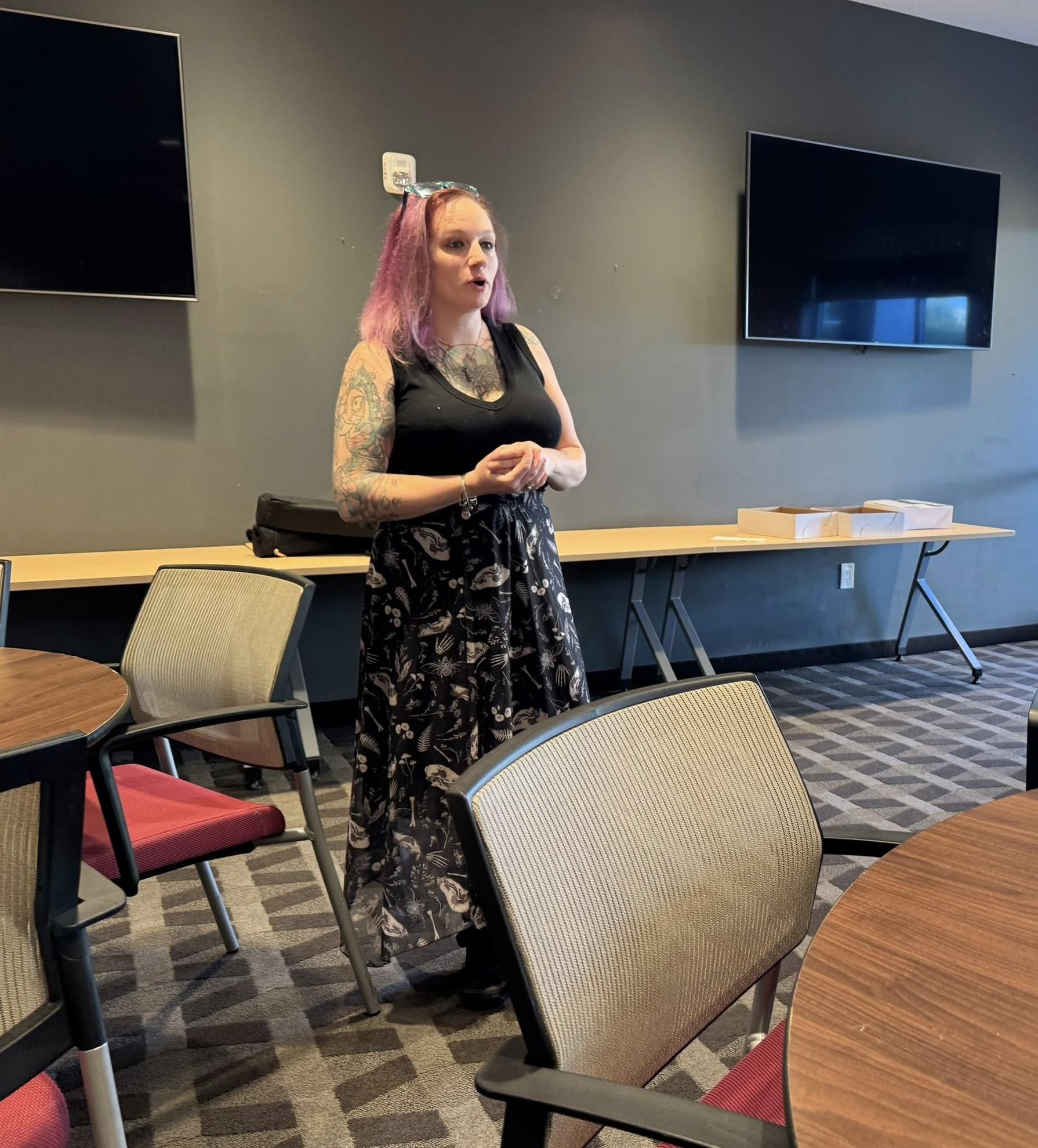 Ashley Angelone of Cleveland Restaurant Group Ashley Angelone, pastry chef, presenting her vision for The Wicked Whisk, a new bakery to open in Solon. She has purple hair, tattoos, and is wearing a black tank top and black skirt with dinosaur bones on it.
