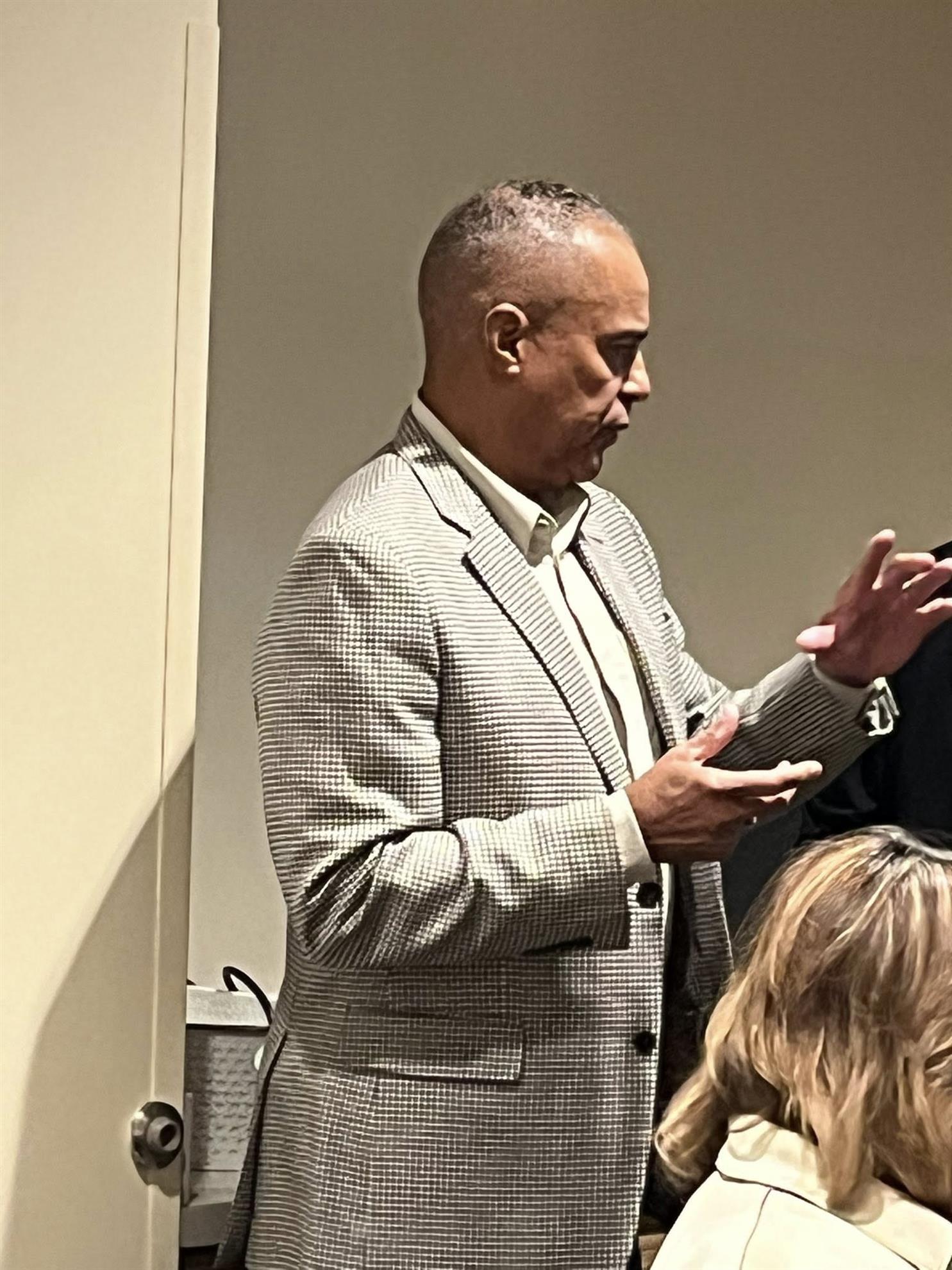 A man with short hair in a greyish and patterned suit is pictured in profile speaking to a room and gesturing with his hands.