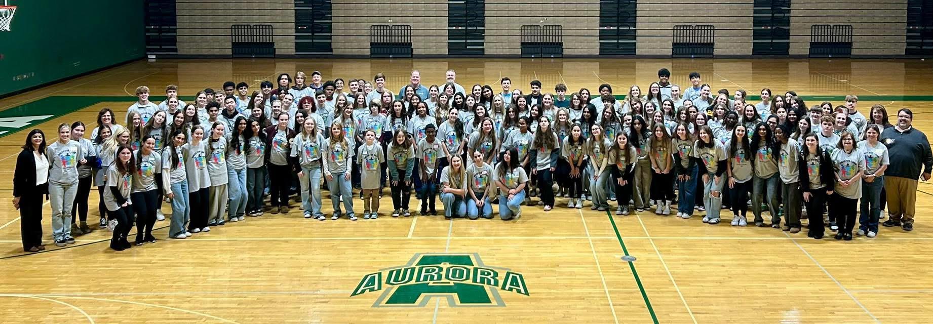 A group of about 100 high school students are pictured in matching grey t-shirts with various adult mentors in a high school gymnasium.