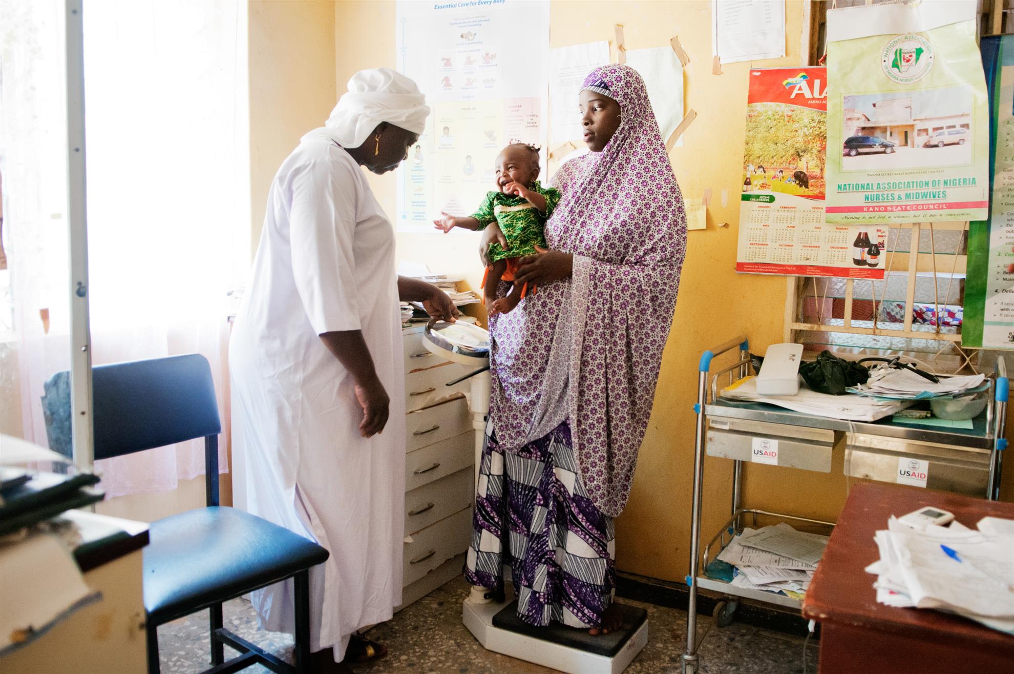 La partera Tamar Okoh (izquierda) pesa a Azima Yahaya y a su hija Aisha en el Hospital General de Sumaila en Sumaila, Estado de Kano (Nigeria). Puesto que no cuenta con una balanza para niños, ella tendrá que calcular el peso de la niña restando el peso de la madre del de la madre con su hija. 4 de septiembre de 2015. El Grupo de Acción Rotaria de Población y Desarrollo (RFPD por sus siglas en inglés) apoya la salud materno-infantil en Nigeria desde hace casi 20 años. En mayo de 2015, cuatro estados nigerianos adoptaron formalmente el proyecto de Control de Calidad en Obstetricia del RFPD, asegurándose así la contínua evaluación y mejora de los estándares y recursos, así como el intercambio de datos. Artículo publicado en la página 54 del número de diciembre de 2015 de la revista