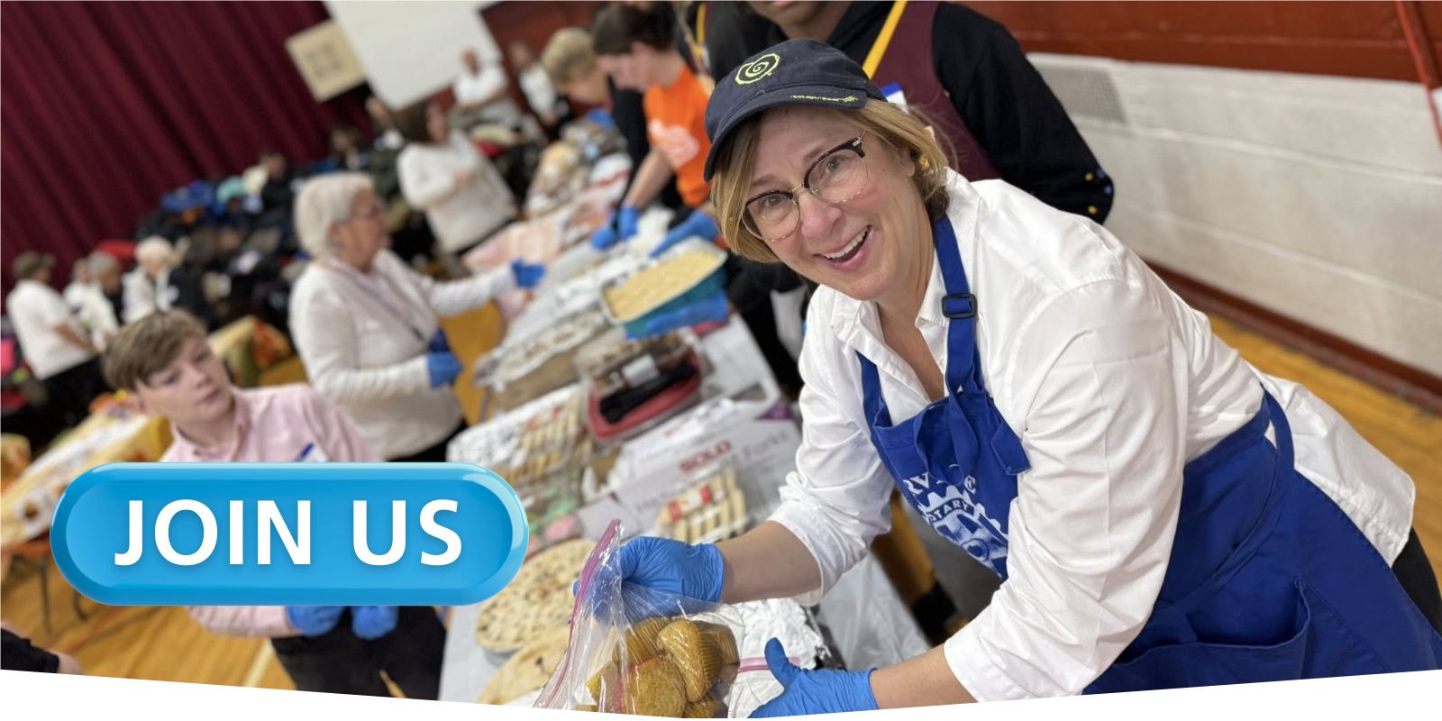 A smiling woman who is a member of Baldwinsville Rotary is wearing a blue apron while serving food in a room filled with people