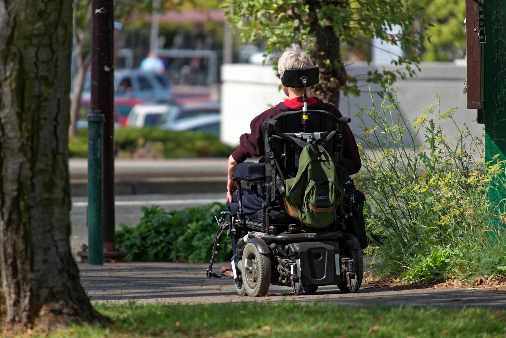 Person using a motorized wheelchair moving independently along an outdoor path, representing long-term mobility, autonomy, and life with a chronic condition.