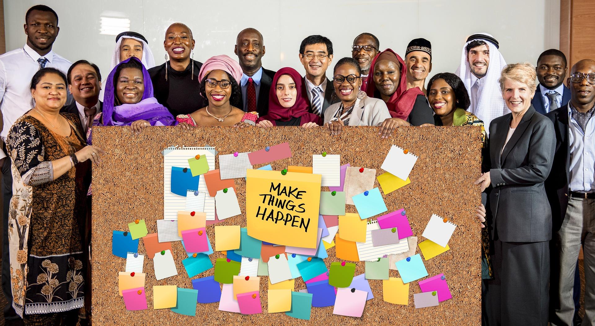 A diverse group of people standing together and smiling behind a large corkboard covered with colorful notes and a central message that reads “Make Things Happen,” symbolizing teamwork, diversity, and collaboration.