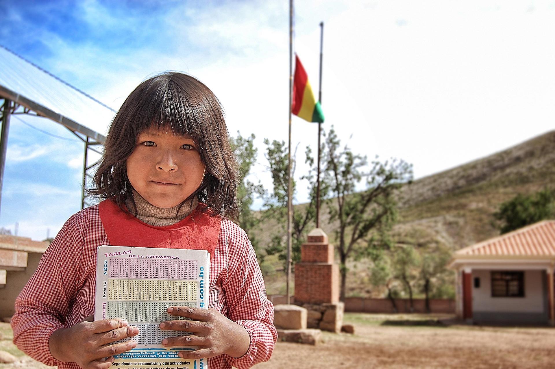 A young student stands outdoors holding a workbook, with a Bolivian flag and a school building in the background — representing access to education and inclusion for all learners.