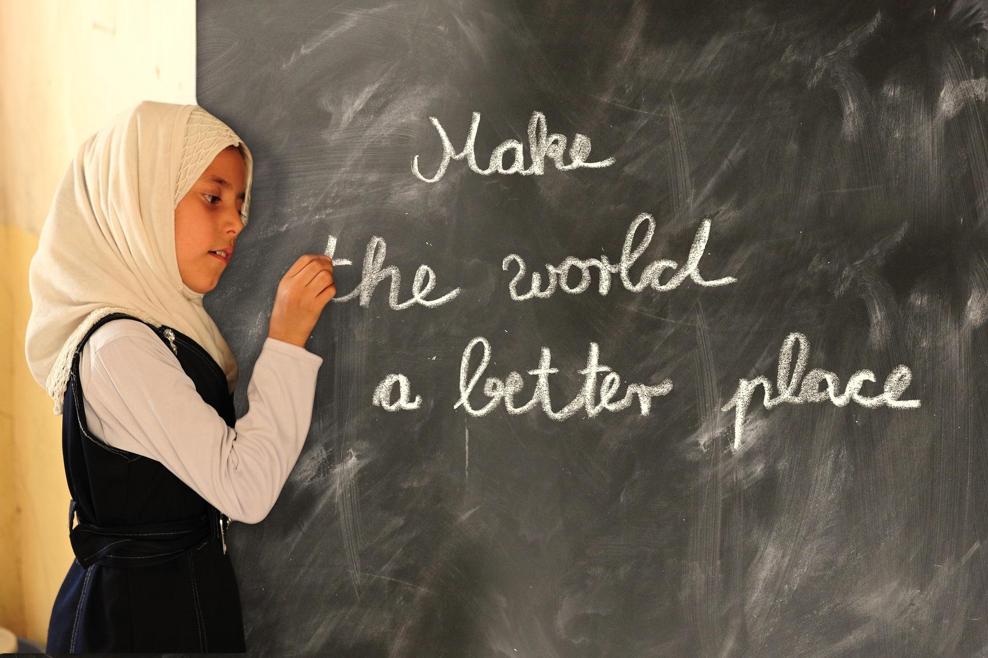 A young girl wearing a light headscarf writes on a chalkboard the words “Make the world a better place,” symbolizing education, hope, and the power of learning to create positive change.