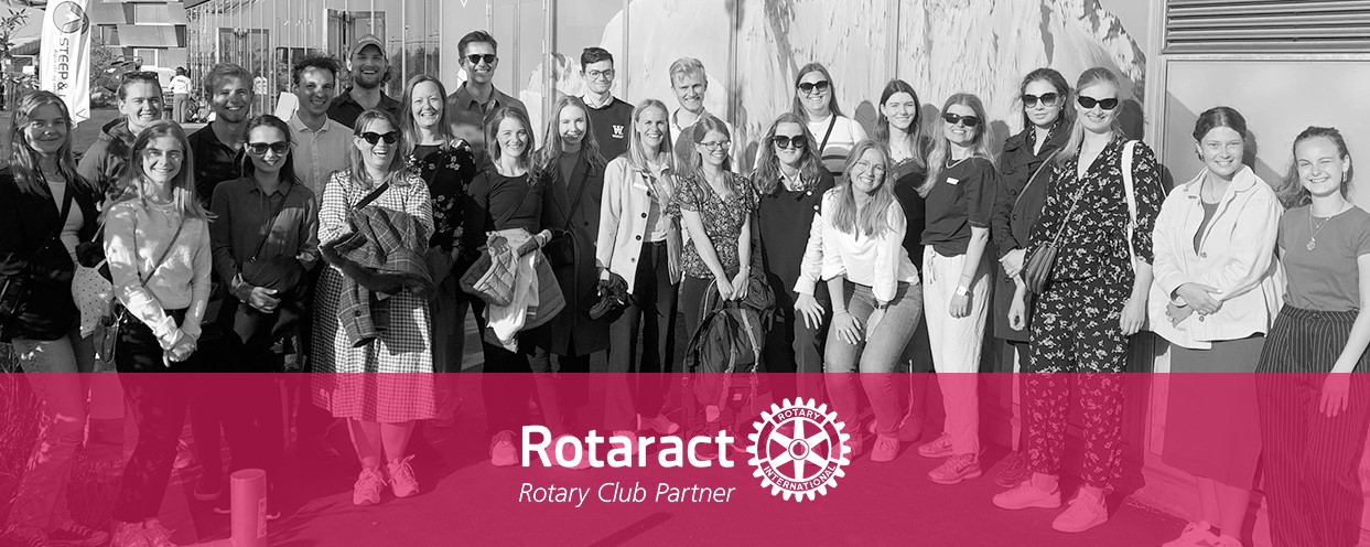 Banner med sort/hvid billede af en masse unge mennesker, der står i solen og smiler til kameraet. I bunden af billedet er der et lyserødt banner med Rotary logo.